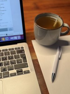 Laptop geöffnet mit Blatt Papier und Stift daneben und einer Tasse Tee.
