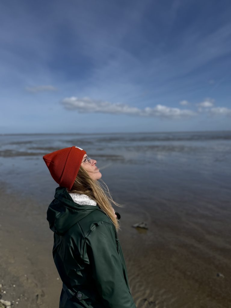 Karen Blömer an der Nordsee mit grünem Regenmantel und roter Mütze.