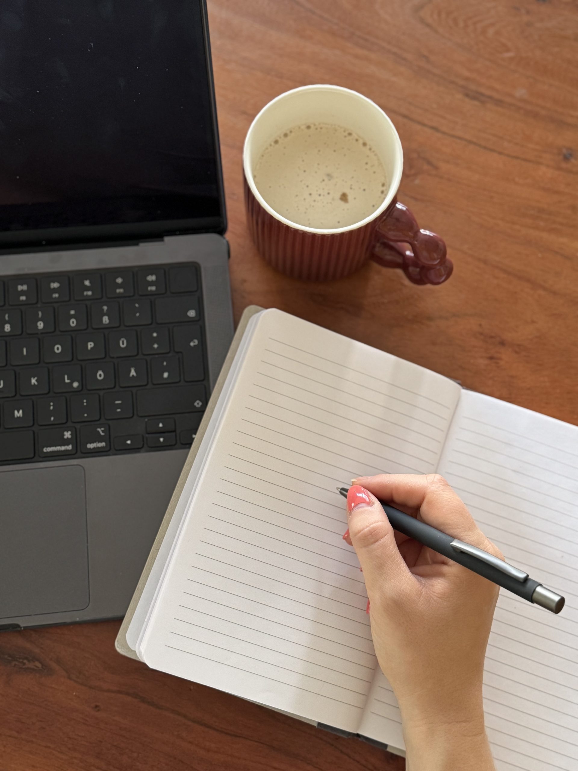 Vogelperspektive: Laptop, Notitzbuch in das ich mit einem Stift schreibe und eine Tasse Kaffe.