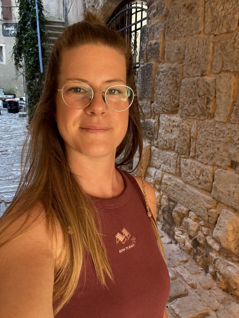 Karen Blömer lächelt in die Kamera, mit roten Top und vor einer Mauer stehend.