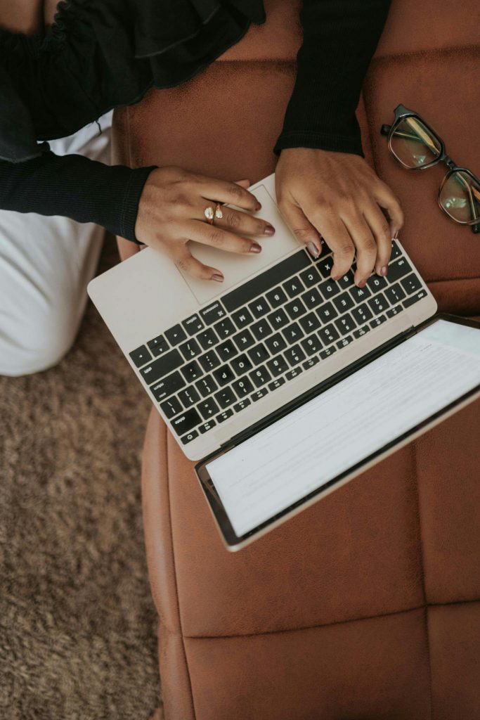Eine Frau, die einen Ring am Finger trägt, tippt auf einem Laptop auf einer braunen Couch mit einer Brille daneben liegend.